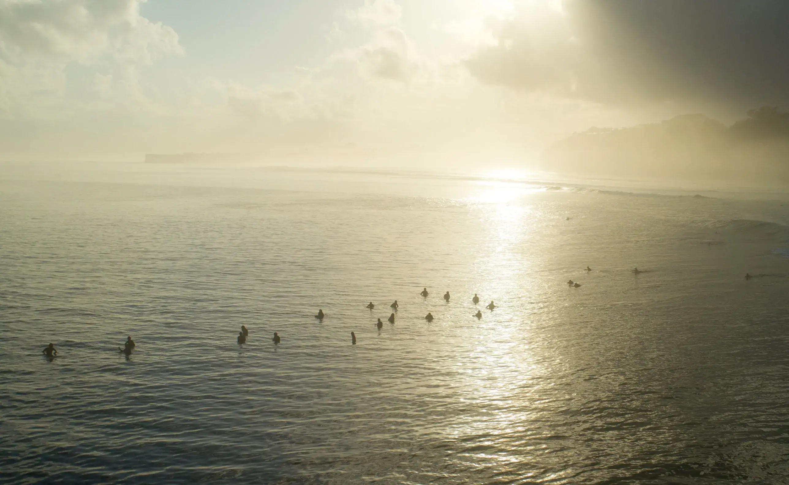 Aerial view of surfers waiting in the lineup on a misty, sunlit morning.